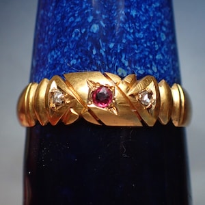 May include: A gold ring featuring a red gemstone and two smaller white gemstones. The ring is displayed against a blue background.