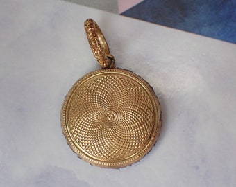 May include: A gold-colored, round pendant with a textured, spiral design on the front. The pendant has a decorative loop at the top. The edges of the pendant have a dark, possibly black, border.