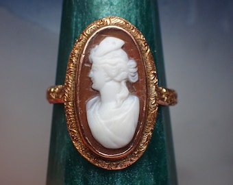 May include: A gold ring with an oval cameo featuring a white profile of a woman with a white hat. The cameo is set in a gold frame with intricate details.