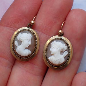 May include: A pair of gold tone earrings with oval shaped cameo pendants featuring white cameo portraits of women.