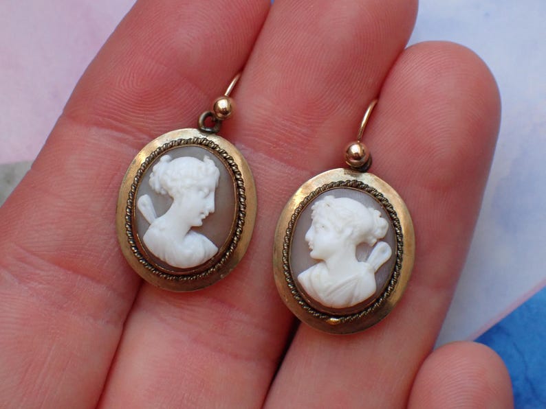 May include: A pair of gold tone earrings with oval shaped cameo pendants featuring white cameo portraits of women.