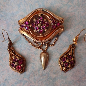 May include: A vintage gold-toned jewelry set featuring a brooch and a pair of earrings, each adorned with vibrant pink gemstones. The brooch has a central design with a teardrop pendant. The earrings have a teardrop shape.