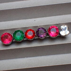 May include: A barrette featuring six colorful gemstones in a row. The gemstones are round, except for the square, clear one at the end. The colors include pink, green, red, purple, and clear. The barrette is set against a gray background.