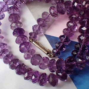 May include: A close-up of a necklace made of faceted purple amethyst beads. The beads are strung together with a silver clasp. The necklace is displayed on a patterned background with shades of blue, white, and pink.