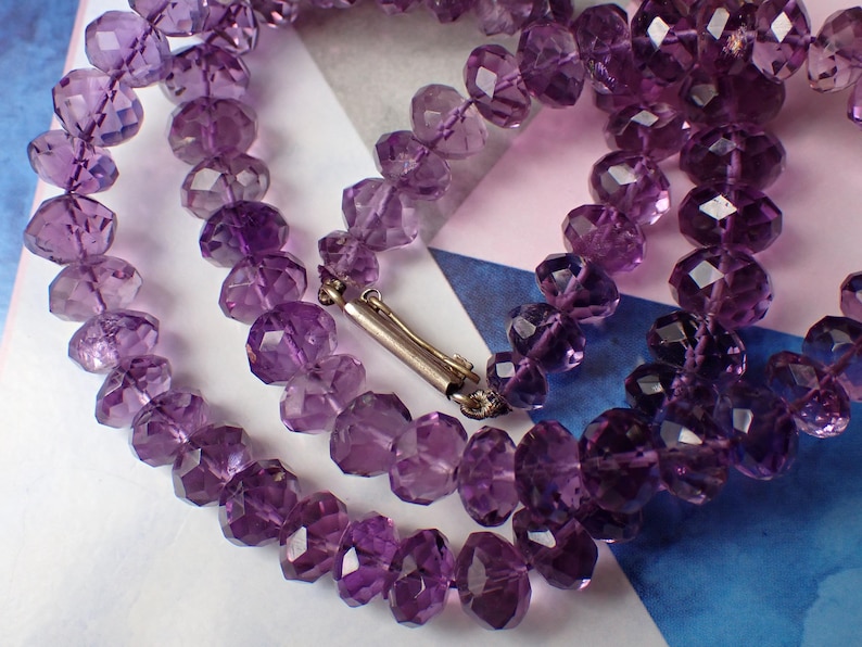 May include: A close-up of a necklace made of faceted purple amethyst beads. The beads are strung together with a silver clasp. The necklace is displayed on a patterned background with shades of blue, white, and pink.
