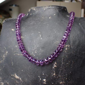 May include: A faceted amethyst bead necklace displayed on a black mannequin. The necklace is a deep purple color and appears to be strung together. The beads are uniform in size and shape. The necklace is a jewelry item.