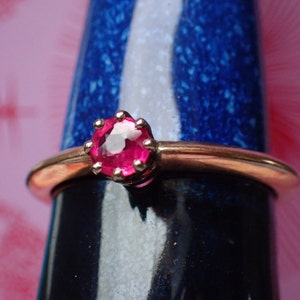 May include: A close-up of a gold ring with a single, round, bright pink gemstone set in a prong setting.