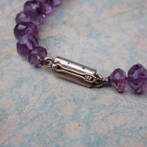 May include: Close-up of a purple amethyst bead necklace with a silver clasp. The faceted beads are a deep violet color and strung together with a silver wire. The clasp is a simple, rectangular design.