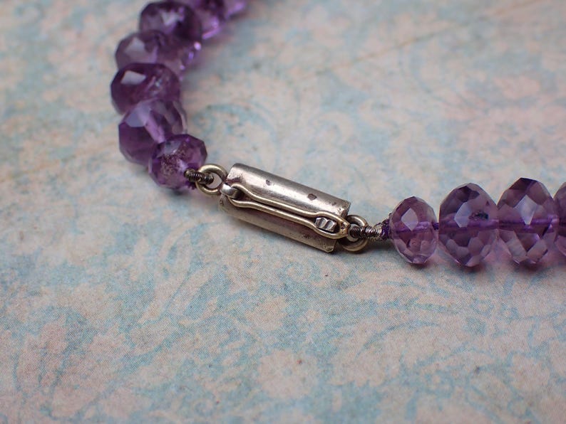 May include: Close-up of a purple amethyst bead necklace with a silver clasp. The faceted beads are a deep violet color and strung together with a silver wire. The clasp is a simple, rectangular design.