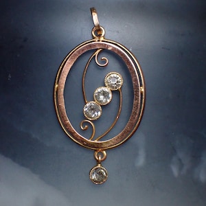 May include: A gold pendant featuring a delicate, oval-shaped frame with a floral design. Four small, clear gemstones are set within the design, and a fifth gemstone hangs from the bottom of the pendant.