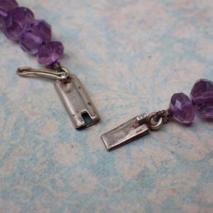 May include: Close-up of a partially unclasped amethyst bead necklace. The necklace features faceted, purple beads and a silver-colored clasp. The background is a light blue and white patterned surface.