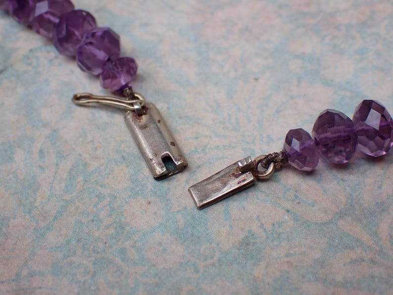 May include: Close-up of a partially unclasped amethyst bead necklace. The necklace features faceted, purple beads and a silver-colored clasp. The background is a light blue and white patterned surface.
