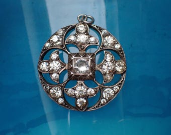 May include: A silver pendant with a circular design featuring a central square with a large clear crystal surrounded by smaller crystals. The pendant has a delicate, intricate design with a floral motif.