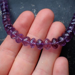 May include: A necklace composed of faceted, deep purple amethyst beads. The beads are strung together, creating a continuous loop. The necklace is displayed against a dark background.