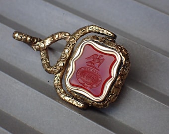 May include: A gold-toned metal swivel fob pendant featuring an ornate, textured frame and a central square-shaped red stone inlay. The stone is engraved with a detailed eagle emblem above a circular crest containing small, intricate text.