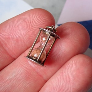 May include: A miniature hourglass pendant crafted from silver-toned metal with a clear glass chamber containing fine golden sand. The small charm features a circular top and bottom frame connected by four vertical pillars and includes a small jump ring.