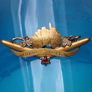 May include: A gold-toned brooch featuring the outline of Australia with the word "AUSTRALIA" engraved. The brooch includes a banner with "GOOD LUCK", a red star, and floral details with pearls. Set against a blue backdrop.