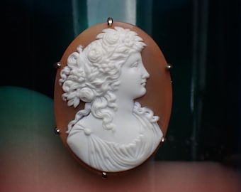 Może przedstawiać: Owalna broszka z kameą przedstawiająca biały relief profilu kobiety na brązowym tle. Kobieta ma kręcone włosy i nosi drapowaną szatę. Broszka jest zabezpieczona srebrnymi szpilkami.