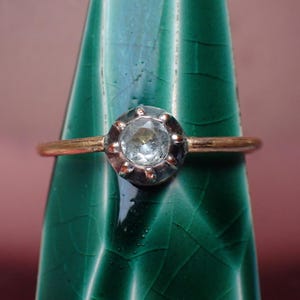 May include: A gold ring with a clear, round gemstone set in a silver-colored bezel. The ring is displayed against a green, textured background with a blurred pink backdrop. The gemstone has a faceted appearance.