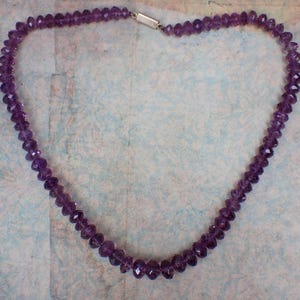 May include: A necklace composed of faceted, deep purple amethyst beads. The beads are strung together, forming a continuous loop. The necklace is secured with a silver-colored clasp. The necklace is displayed against a light blue background.