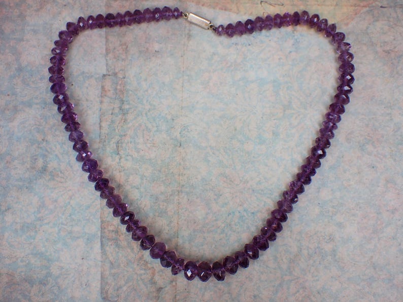 May include: A necklace composed of faceted, deep purple amethyst beads. The beads are strung together, forming a continuous loop. The necklace is secured with a silver-colored clasp. The necklace is displayed against a light blue background.