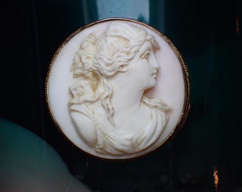 Puede incluir: Broche de camafeo redondo con un perfil tallado de una mujer con cabello suelto y ropa drapeada. El relieve de color marfil se encuentra sobre un fondo blanco, enmarcado por un borde de metal dorado. Una pieza de joyería clásica.