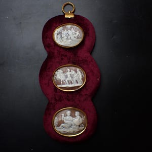 May include: A burgundy velvet wall hanging with three oval cameos. Each cameo, set in a gold-coloured frame, depicts a classical relief scene. A gold ring at the top allows for hanging. The overall design is ornate.