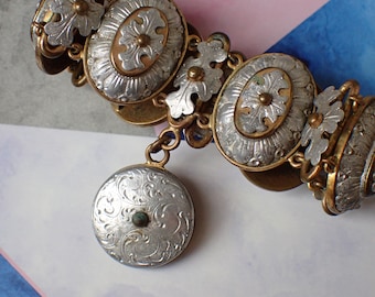 May include: A silver and gold bracelet with a decorative design. The bracelet is made up of several oval-shaped charms with intricate floral patterns. A smaller round charm hangs from the bracelet.