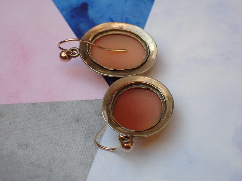 May include: A pair of antique gold tone earrings with a cameo design. The cameos are oval shaped and feature a pink colored design.