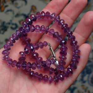 May include: A faceted amethyst necklace with a silver clasp is displayed in an open hand. The necklace is composed of small, round, purple gemstones. The background is a patterned fabric.