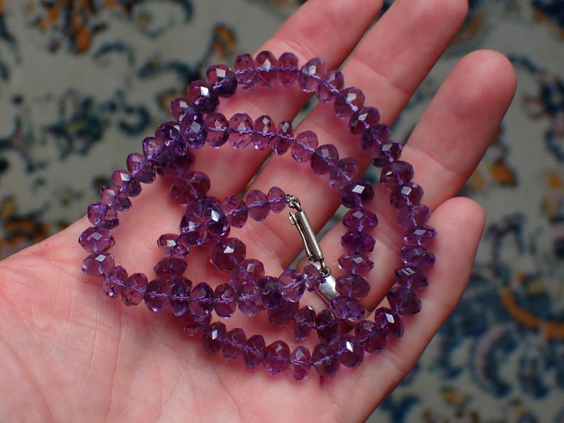 May include: A faceted amethyst necklace with a silver clasp is displayed in an open hand. The necklace is composed of small, round, purple gemstones. The background is a patterned fabric.