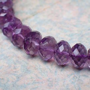 May include: A strand of faceted amethyst beads. The beads are a deep purple color and have a geometric, multi-faceted design. The beads are strung together on a thin, purple string. The background is a light blue and white floral pattern.