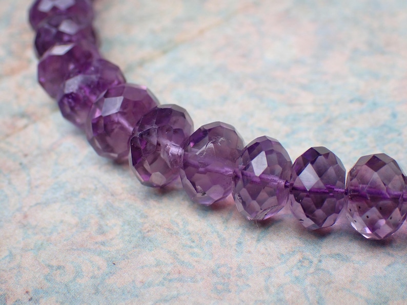 May include: A strand of faceted amethyst beads. The beads are a deep purple color and have a geometric, multi-faceted design. The beads are strung together on a thin, purple string. The background is a light blue and white floral pattern.