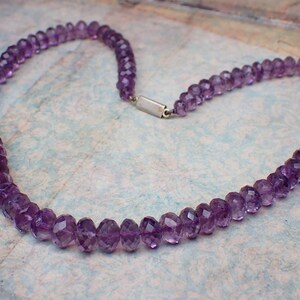 May include: A necklace composed of faceted, deep purple amethyst beads. The beads are strung together, forming a continuous loop with a silver-colored clasp. The necklace is displayed on a patterned surface.