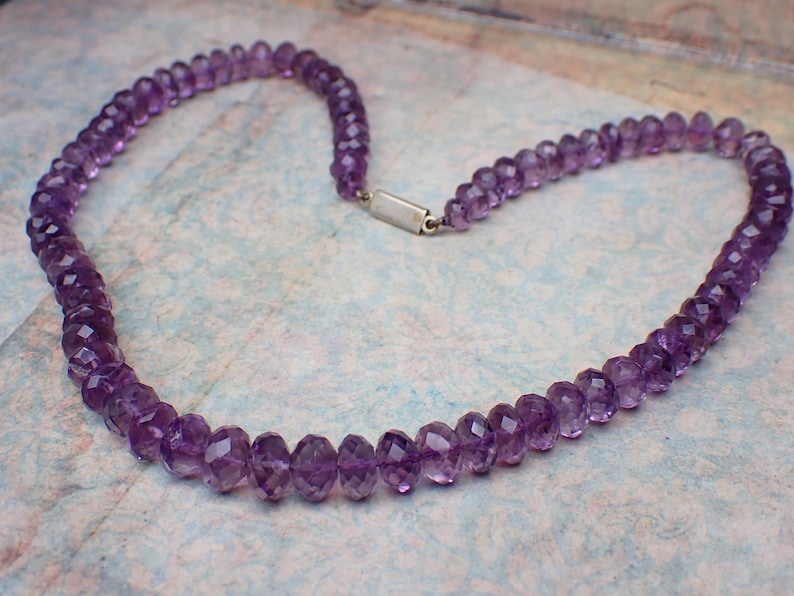 May include: A necklace composed of faceted, deep purple amethyst beads. The beads are strung together, forming a continuous loop with a silver-colored clasp. The necklace is displayed on a patterned surface.