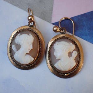 May include: A pair of gold tone earrings with cameo portraits of women. The cameos are set in oval frames with a rope-like border.