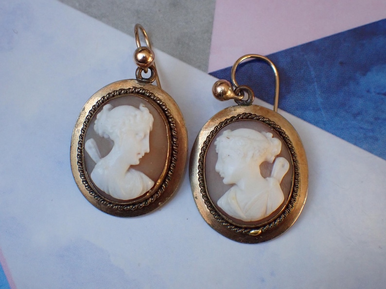 May include: A pair of gold tone earrings with cameo portraits of women. The cameos are set in oval frames with a rope-like border.