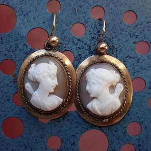 May include: A pair of gold tone earrings with oval shaped cameo portraits of women. The cameos are white and the earrings have a rope-like design around the edges.