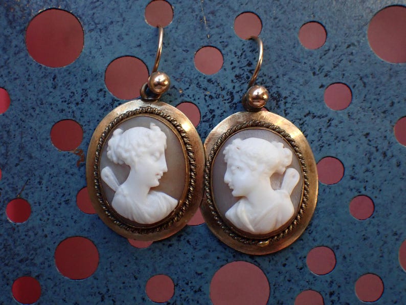 May include: A pair of gold tone earrings with oval shaped cameo portraits of women. The cameos are white and the earrings have a rope-like design around the edges.