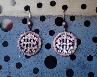 May include: A pair of silver-toned earrings. Each earring features a circular pendant with an ornate, stylized monogram within a decorative border. The earrings have a simple hook design.