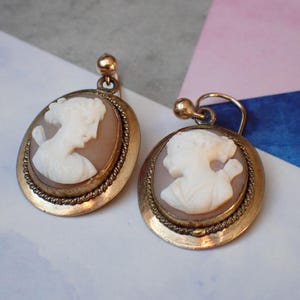 May include: A pair of gold tone earrings with cameo portraits of women. The cameos are set in a round, gold tone frame with a rope-like border.