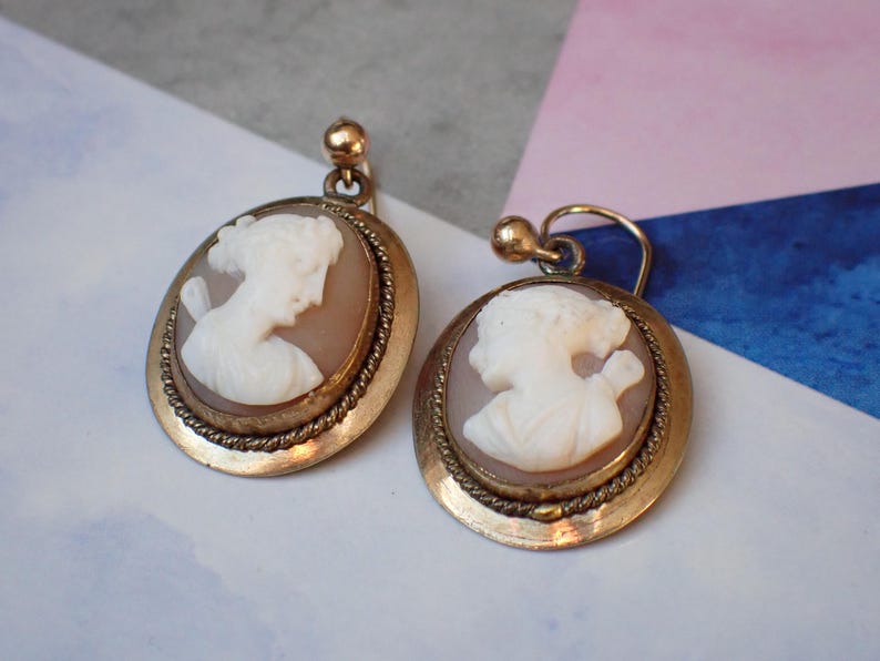 May include: A pair of gold tone earrings with cameo portraits of women. The cameos are set in a round, gold tone frame with a rope-like border.