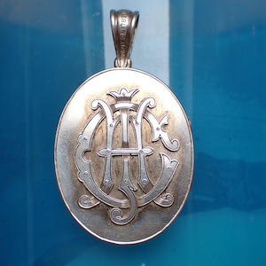 May include: Oval silver pendant with a monogram design. The pendant features the letters "A" and "C" intertwined, topped with a crown. The bail is marked "STERLING SILVER".