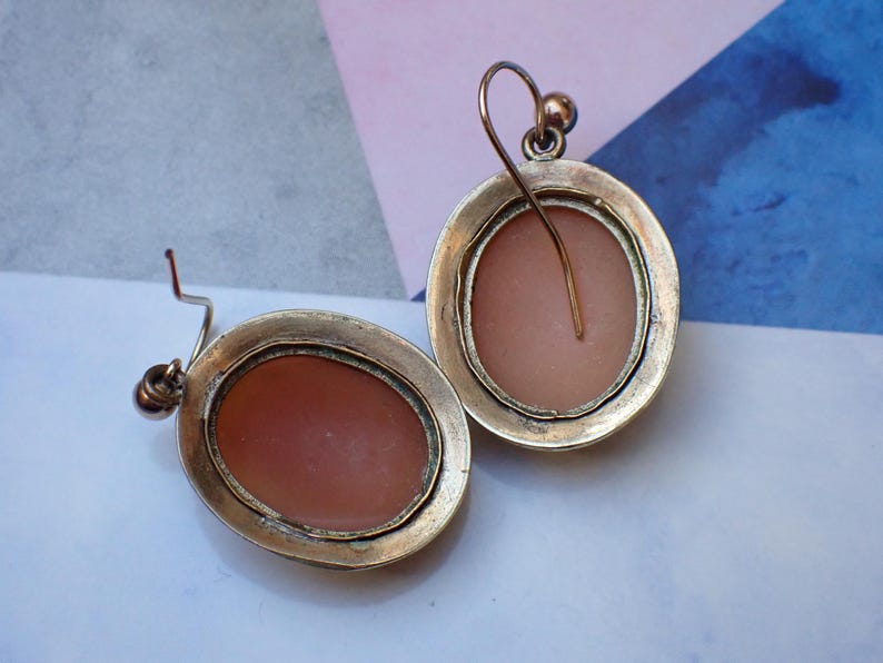 May include: A pair of silver tone earrings with a cameo design. The cameos are oval shaped and have a pink background.