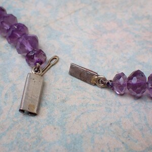 May include: Close-up of a purple amethyst bead necklace with a silver clasp. The faceted beads are a deep violet color and strung together. The clasp is open, showing the necklace's construction.