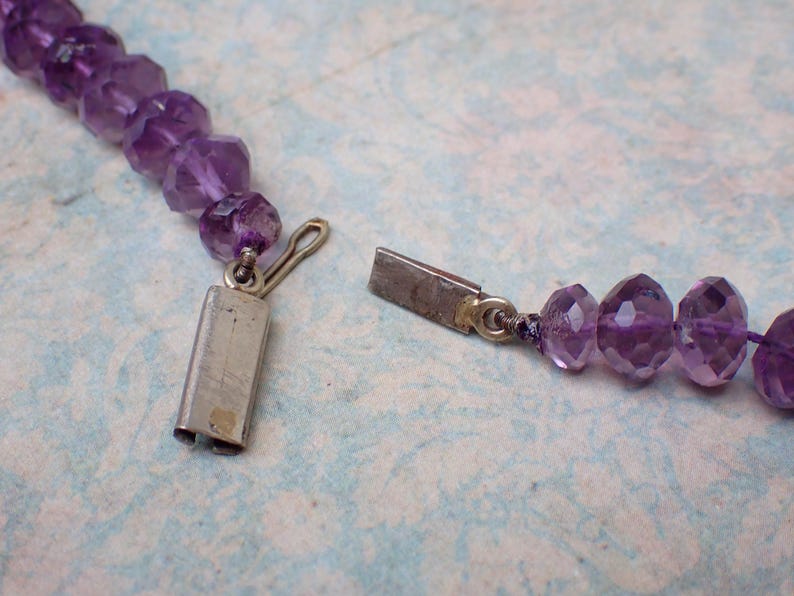 May include: Close-up of a purple amethyst bead necklace with a silver clasp. The faceted beads are a deep violet color and strung together. The clasp is open, showing the necklace's construction.