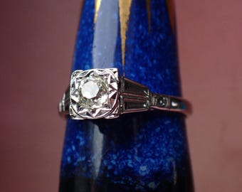 May include: A silver engagement ring with a round, clear diamond set in a geometric design. The ring is on a blue background.