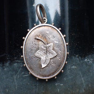 Peut inclure: Pendentif ovale en argent avec un motif de feuille. Le pendentif présente une feuille détaillée au centre, entourée de gravures florales délicates. La bordure est ornée de petits points en relief. Une boucle est en haut pour une chaîne.