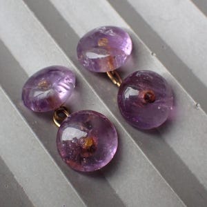 May include: Four amethyst cufflinks with gold-coloured findings. The cufflinks are a translucent purple colour with a rounded shape. The background is a light grey, textured surface.
