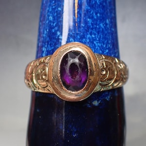 May include: A gold-toned ring with an oval, deep purple gemstone. The ring's band features intricate floral and scrollwork designs. The gemstone is set in a gold bezel, creating a classic and elegant look.